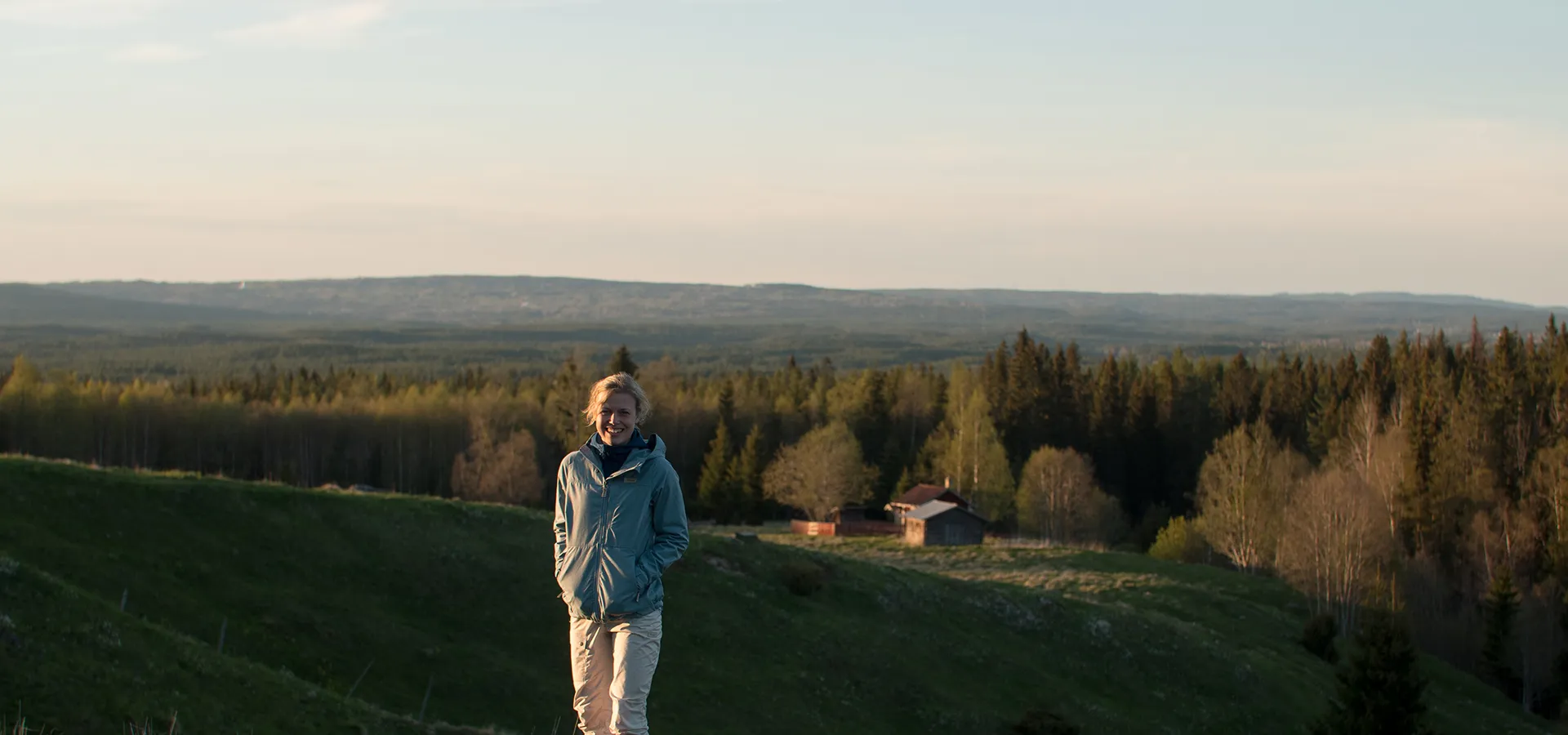 En kvinna står i ett öppet landskap med utsikt över skog och berg.