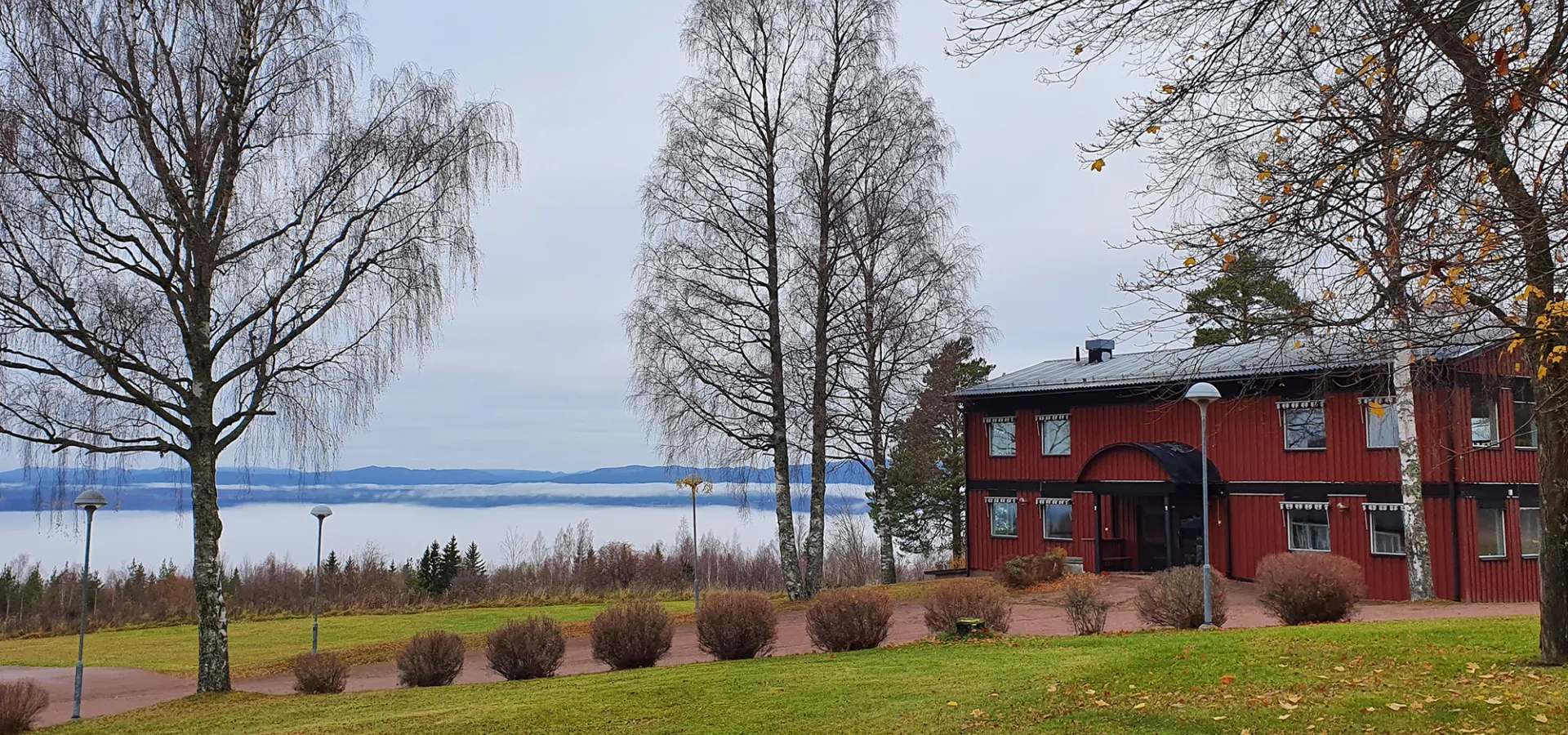 Ett rött hus bredvid kala träd och en grön gräsmatta. En stor sjö i bakgrunden.