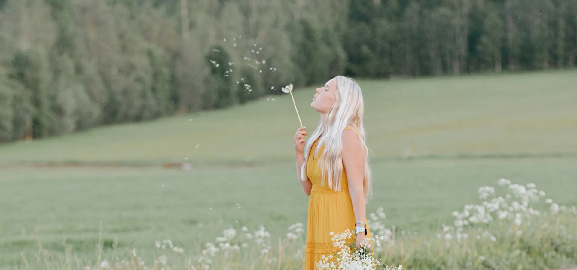 Stina Jansson på en blomsteräng, blåser på en utblommad maskros.