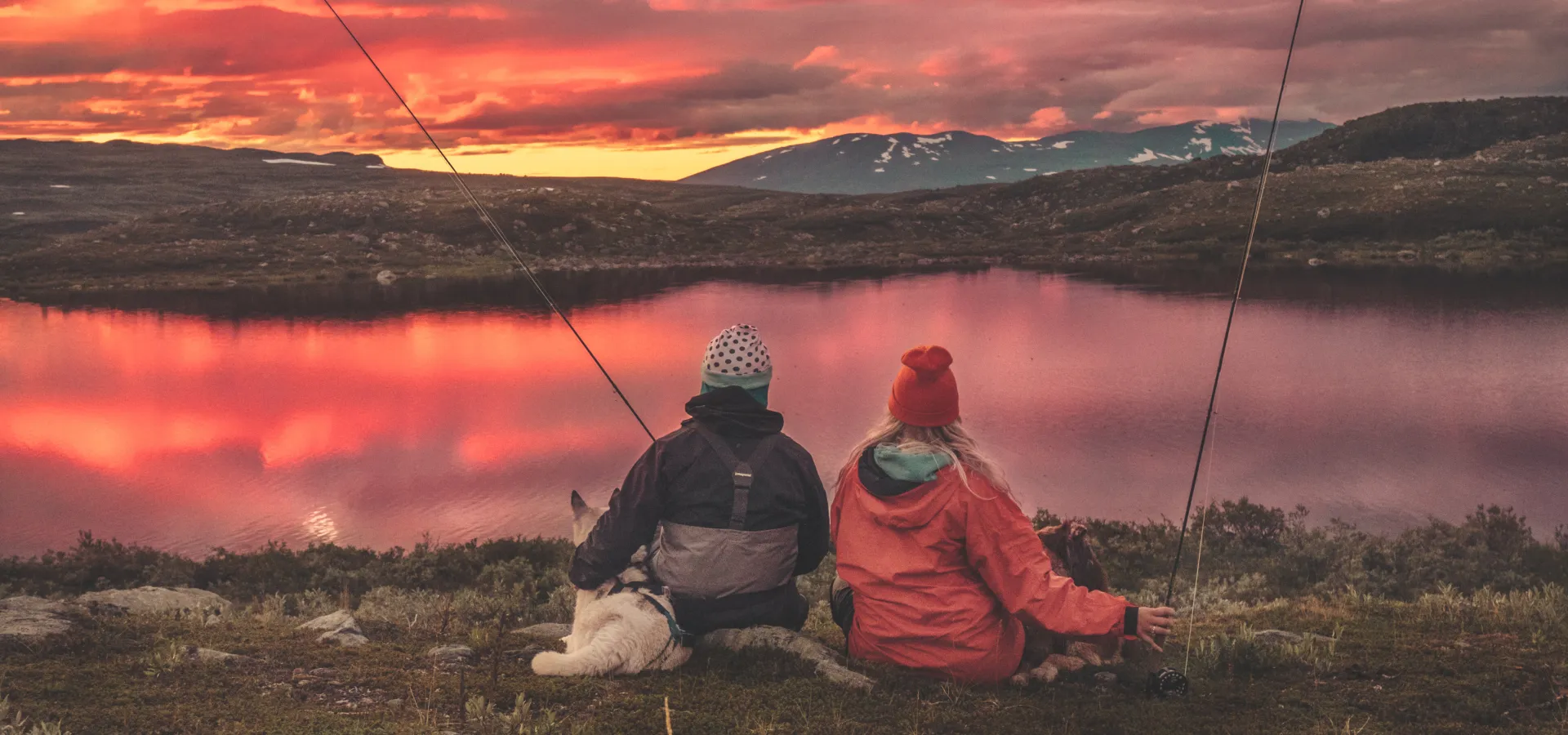 Två personer och en hund sitter vid sjö i fjällen.