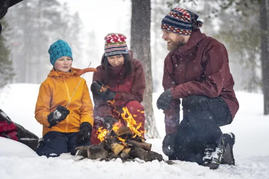 Familj värmer sig runt en öppen eld i snörikt landskap.