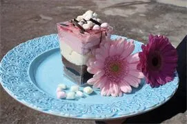 A pastry in three layers, stands on a turquoise dish, a pink and a purple flower lies on the dish.