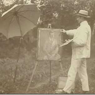 A man standing in a meadow holding a painting resting on an easel. 
