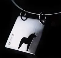 Ett fyrkantigt silversmycke med en utskuren dalahäst, hänger i två öglor i en silvervajer.