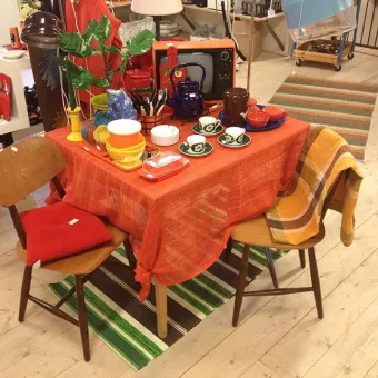 Interiörbild från butiken, ett bord med orange duk, på bordet koppar och diverse varor, randig matta på golvet.