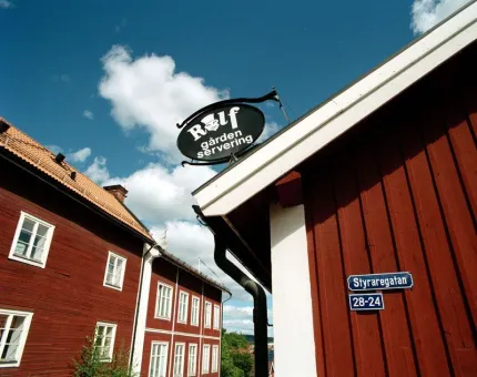 En röd husknut, på väggen en blå skylt Styraregatan, på taket en svart skylt Rolfgården servering, röda trähus på andre sidan gatan.