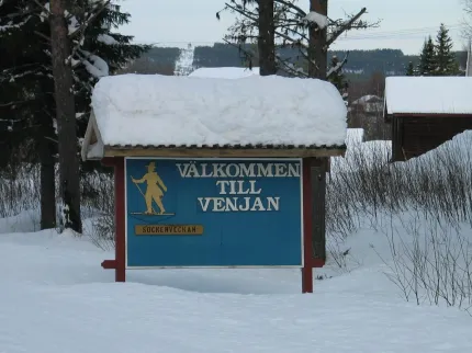 Welcome sign, snow-covered terrain, forest and mountains in the background.