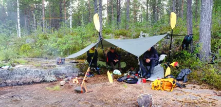 Three people in an open tent in the forest. Their luggage is on the ground.