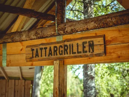 Träskylt med inhuggna bokstäver med namnet Tattargrillen under ett tak.