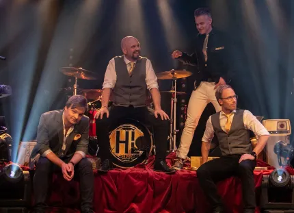 Fyra män på en scen, två sitter på scenkanten, en sitter på en trumma och en står, ett trumset i bakgrunden.