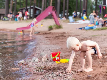 En pojke som leker i sanden i vattenbrynet, röd rutschkana i bakgrunden.